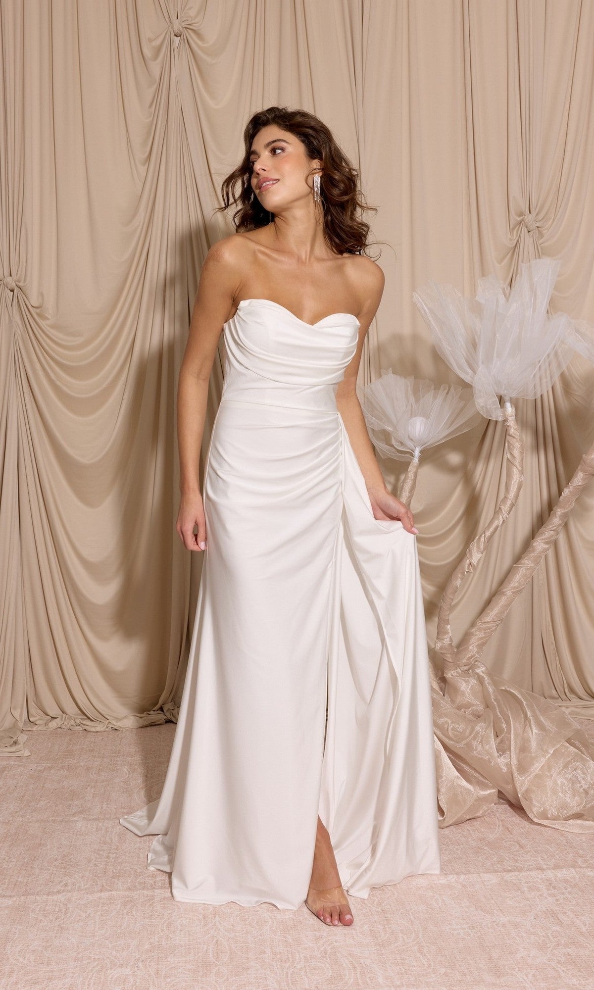 A woman in the off white Long Prom Dress: Eureka EK178 stands barefoot on a light rug, holding one side of her gown, with beige draped curtains and white decorative branches in the background.